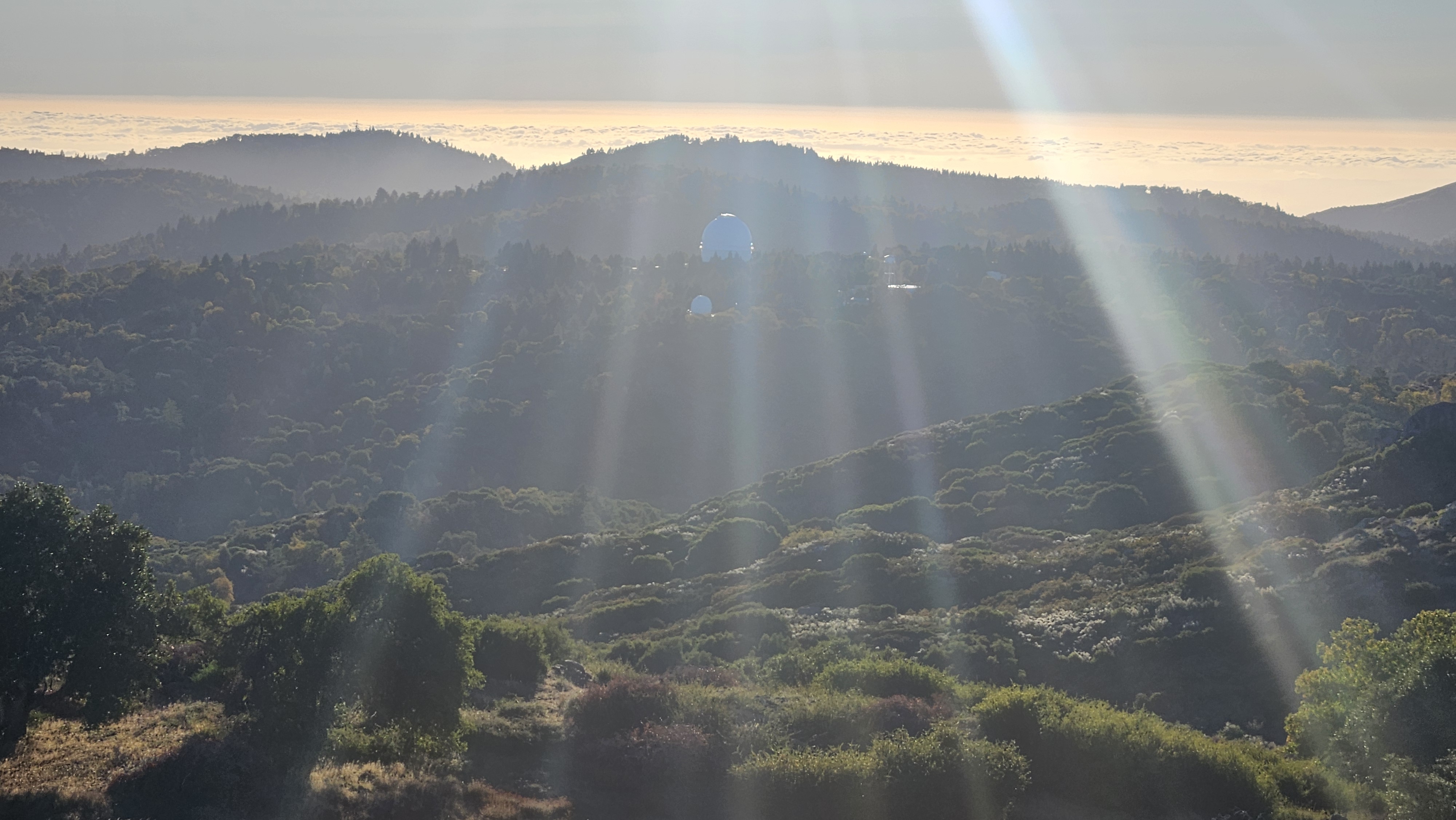 Palomar Observatory domes tucked into the ridges at sunset, marine layer banked behind, sun breaking through